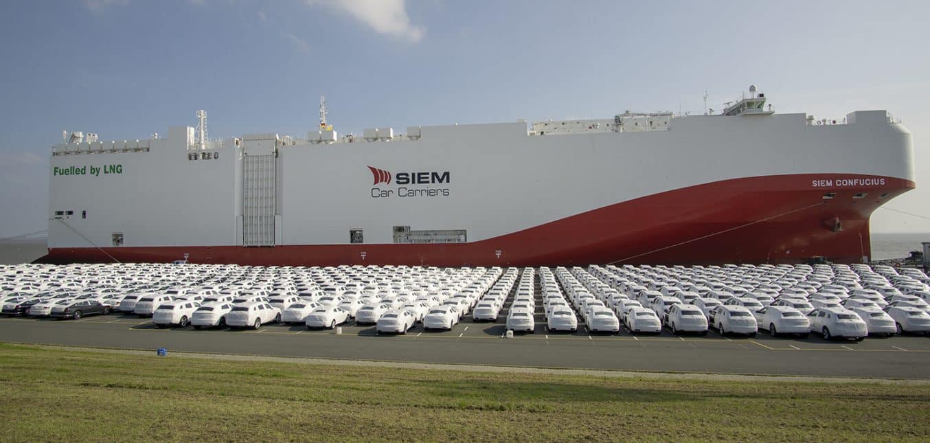A large red and white ship docked next to a row of new cars